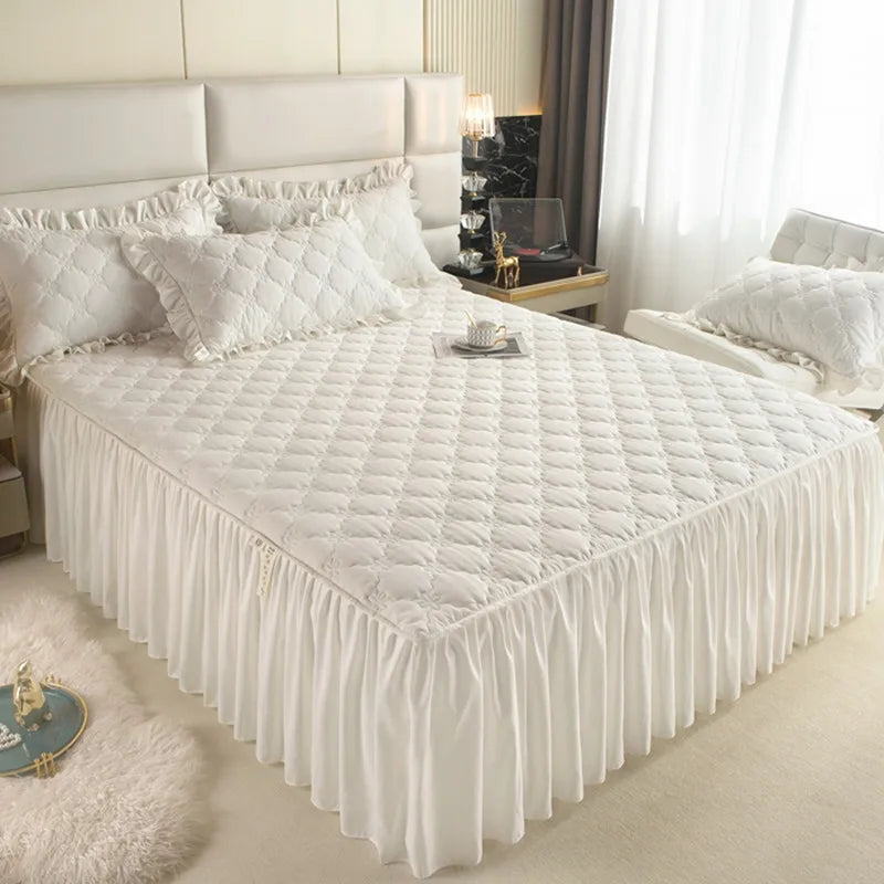 A white quilted bed skirt cover with a gathered bottom is displayed on a bed in a bright bedroom.