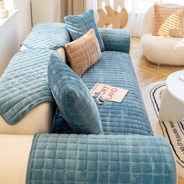 A teal velvet sofa protection cover with quilted cushions and a tan accent pillow on a light-colored sofa.