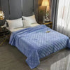 A plush velvet quilted blue bed cover blanket spread on a bed with white pillows and dark gray curtains.