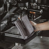 A hand is holding a gray microfiber cleaning towel over the dispensing nozzle of a black coffee machine.
