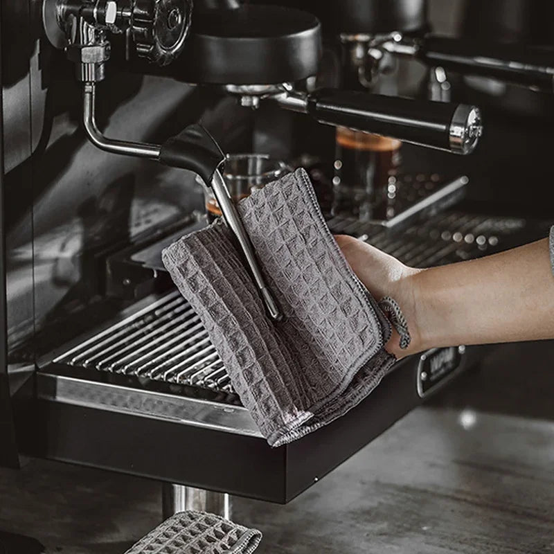 A hand is holding a gray microfiber cleaning towel over the dispensing nozzle of a black coffee machine.