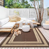 A brown and beige reversible outdoor patio rug placed on a wooden deck with white furniture and greenery in the background.