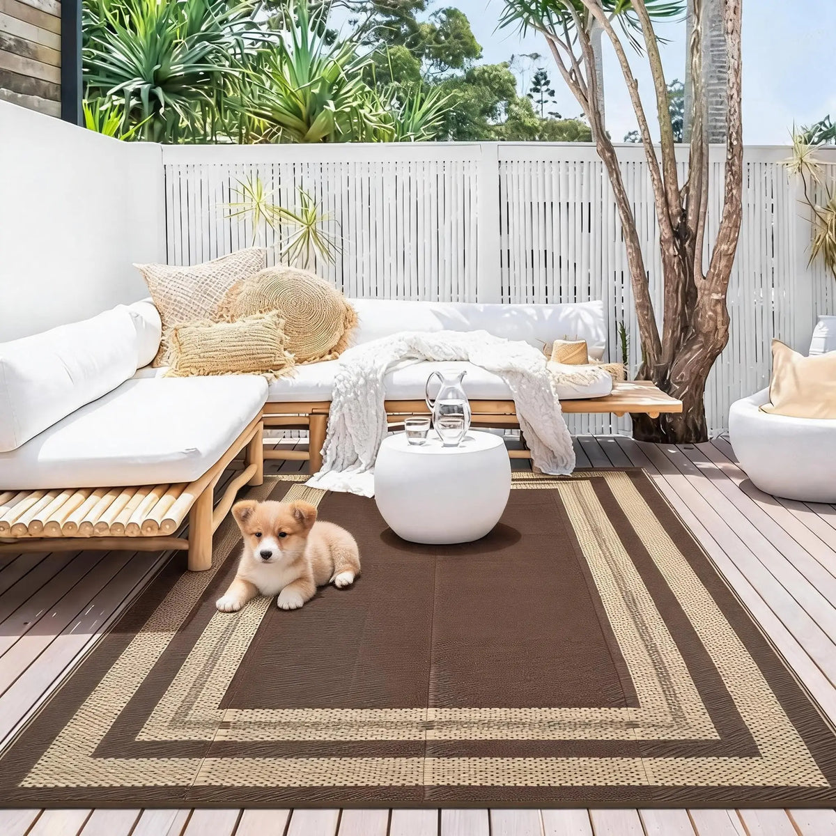 A brown and beige outdoor patio rug is placed on a wooden deck with a white sofa and greenery in the background.