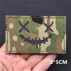 A rectangular tactical patch featuring a camouflage pattern with crossed eyes and a grinning skull face