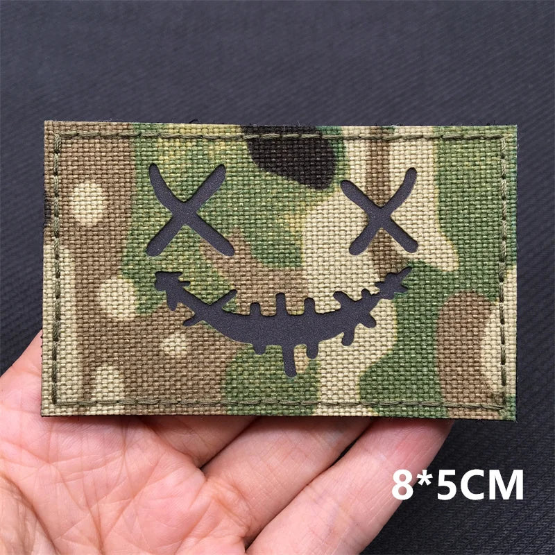 A rectangular tactical patch featuring a camouflage pattern with crossed eyes and a grinning skull face