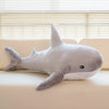 A gray giant shark plush toy lying on a light-colored sofa in front of horizontal blinds.