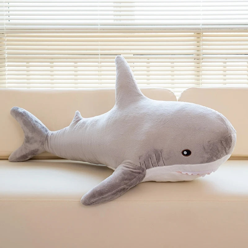 A gray giant shark plush toy lying on a light-colored sofa in front of horizontal blinds.
