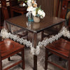 A dark wood dining table covered with a sheer lace embroidered PVC tablecloth and decorated with flowers.