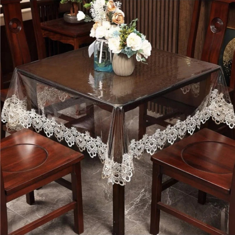 A dark wood dining table covered with a sheer lace embroidered PVC tablecloth and decorated with flowers.
