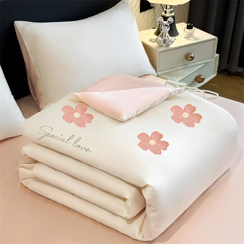 A white embroidered cooling summer quilt with pink floral designs and a matching pink pillow is displayed on a light pink sur