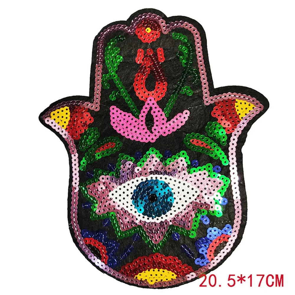 A black iron-on patch featuring a pink and green design with a central blue eye made of sequins.