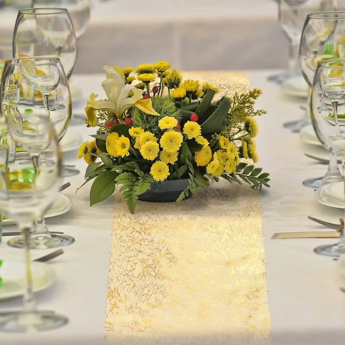 A metallic gold table runner is placed on a white tablecloth, featuring a vibrant bouquet of yellow flowers.