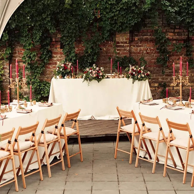 A long white satin tablecloth is set up outdoors against a brick wall with greenery, ready for dining or party decor.