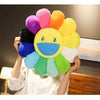 A hand holds a colorful plush sunflower cushion with a smiling face, featuring petals in shades of pink, orange, yellow, and 
