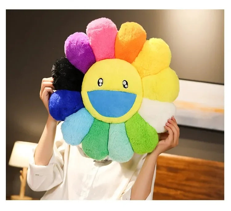 A hand holds a colorful plush sunflower cushion with a smiling face, featuring petals in shades of pink, orange, yellow, and 