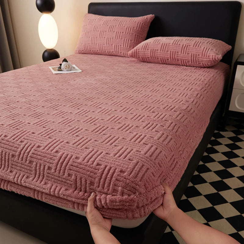A close-up of a pink plush jacquard mattress cover on a bed with dark bedding and patterned flooring.