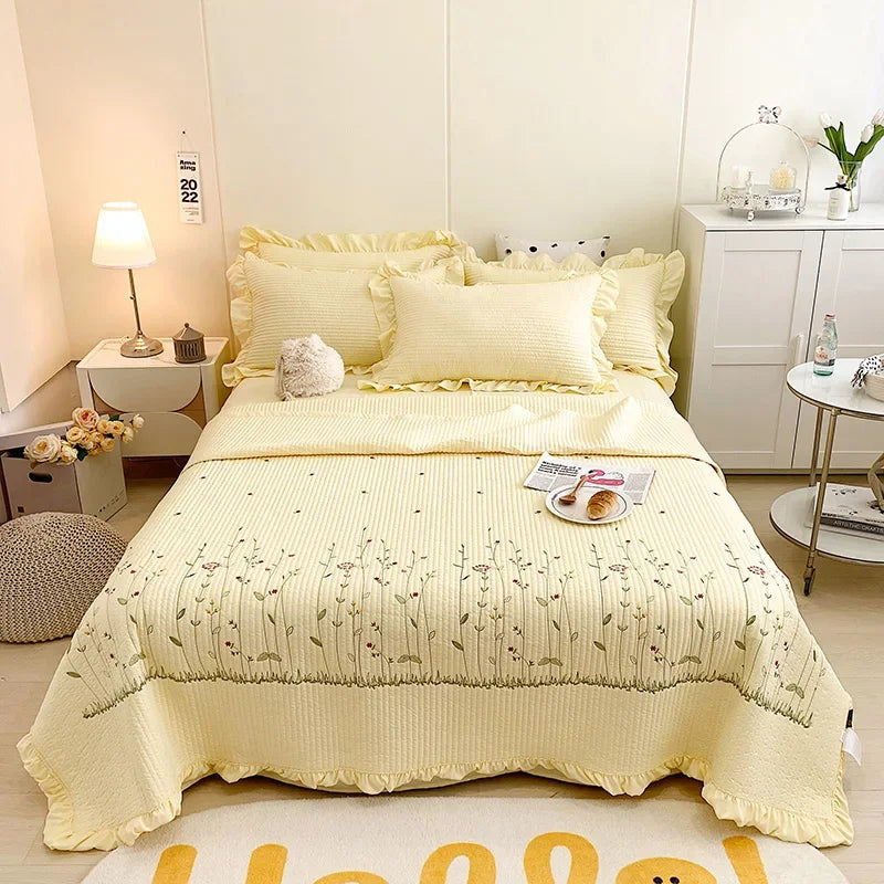 A yellow floral embroidery quilt bedspread set displayed on a bed with white linens and light-colored walls.