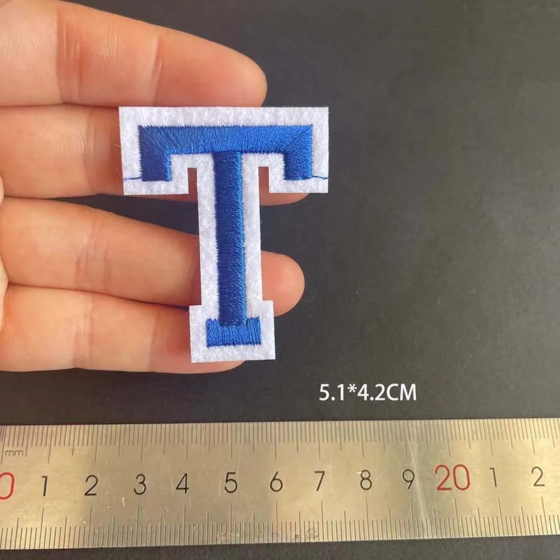 A hand holds a blue alphabet embroidery iron-on patch shaped like the letter T, measured against a ruler.