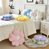 A collection of soft, pastel-colored plush daisy flower throw pillows arranged on a white bed and a bedside table.