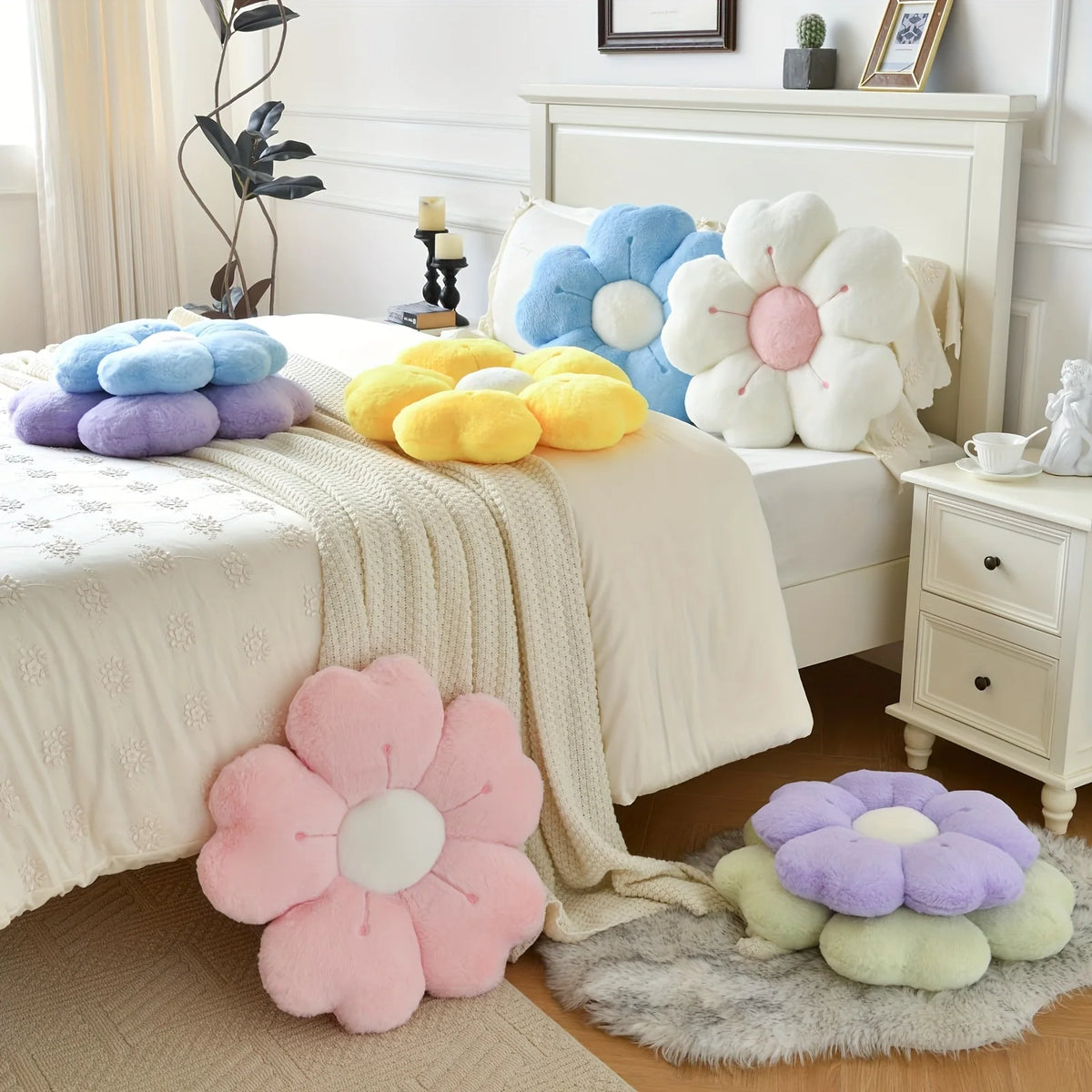 A collection of soft, pastel-colored plush daisy flower throw pillows arranged on a white bed and a bedside table.