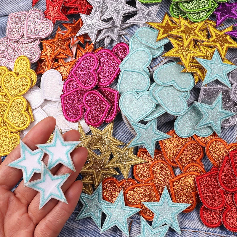 A hand holds a collection of glitter iron-on embroidered love heart and star patches in various colors like pink, gold, silve