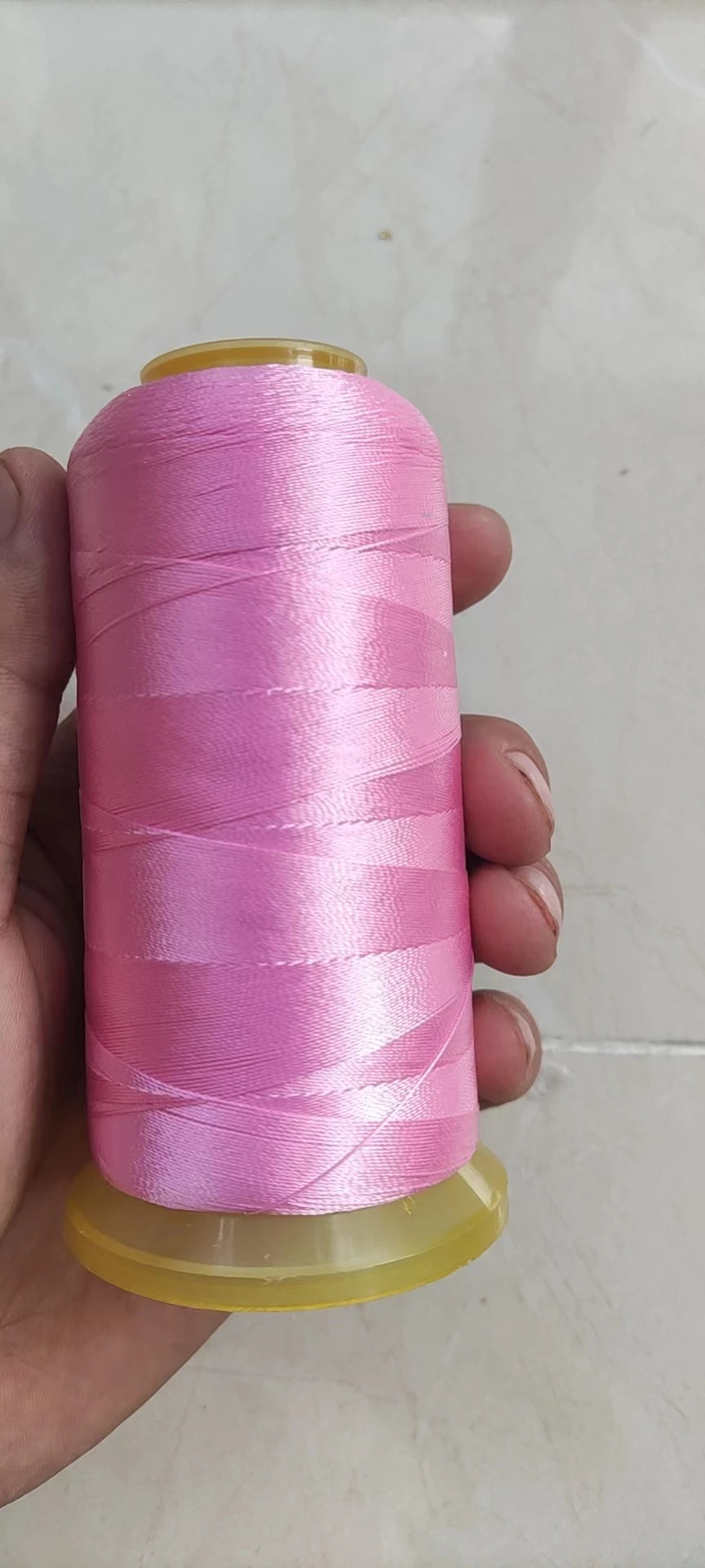 A hand holds a spool of bright pink high tenacity polyester sewing thread with a yellow plastic core.
