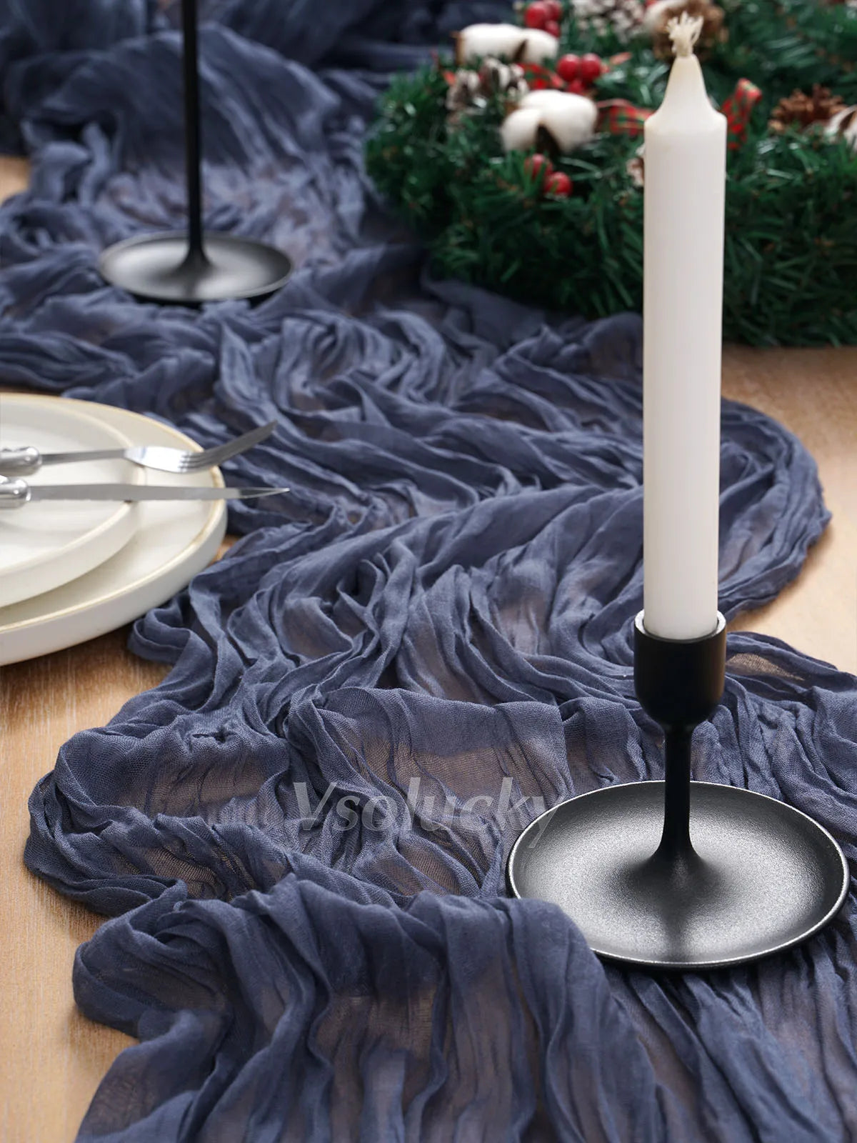 A dark blue crinkled cheesecloth table runner draped over a wooden surface with a white candle and silver cutlery.