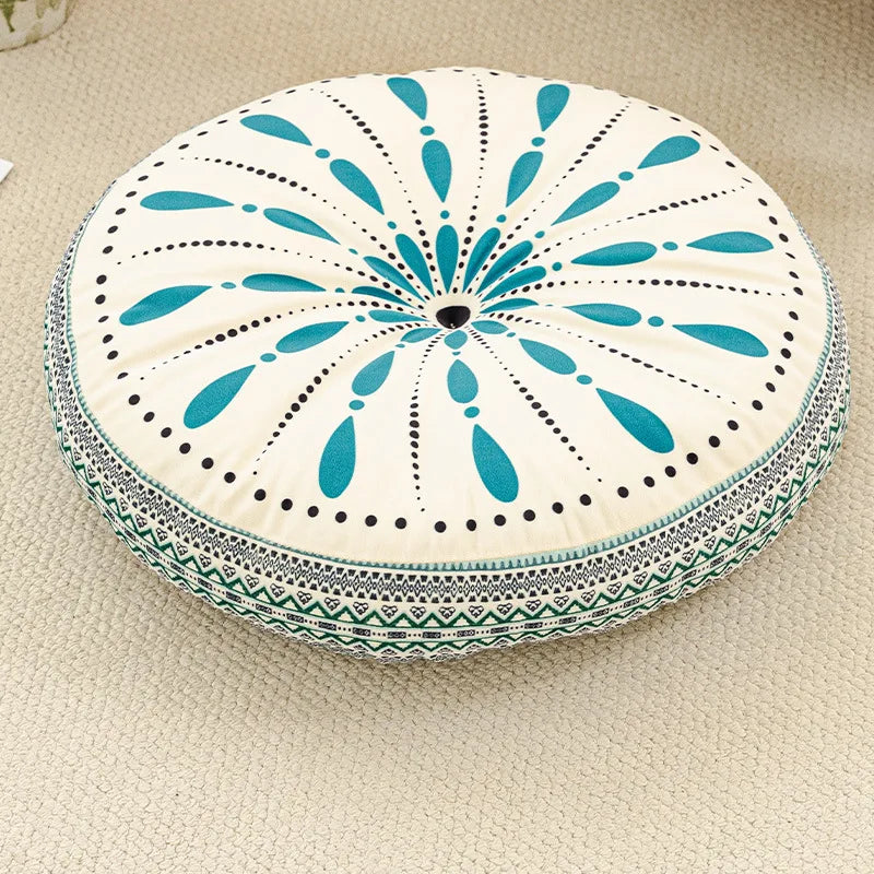 A round cushion with a white and teal floral pattern featuring radiating petals and an embroidered border.