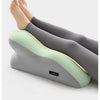 A pair of legs and feet resting on a gray ergonomic pillow with a light green insert, used for stomach sleeping.