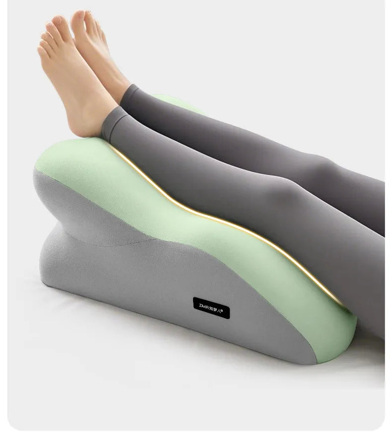 A pair of legs and feet resting on a gray ergonomic pillow with a light green insert, used for stomach sleeping.