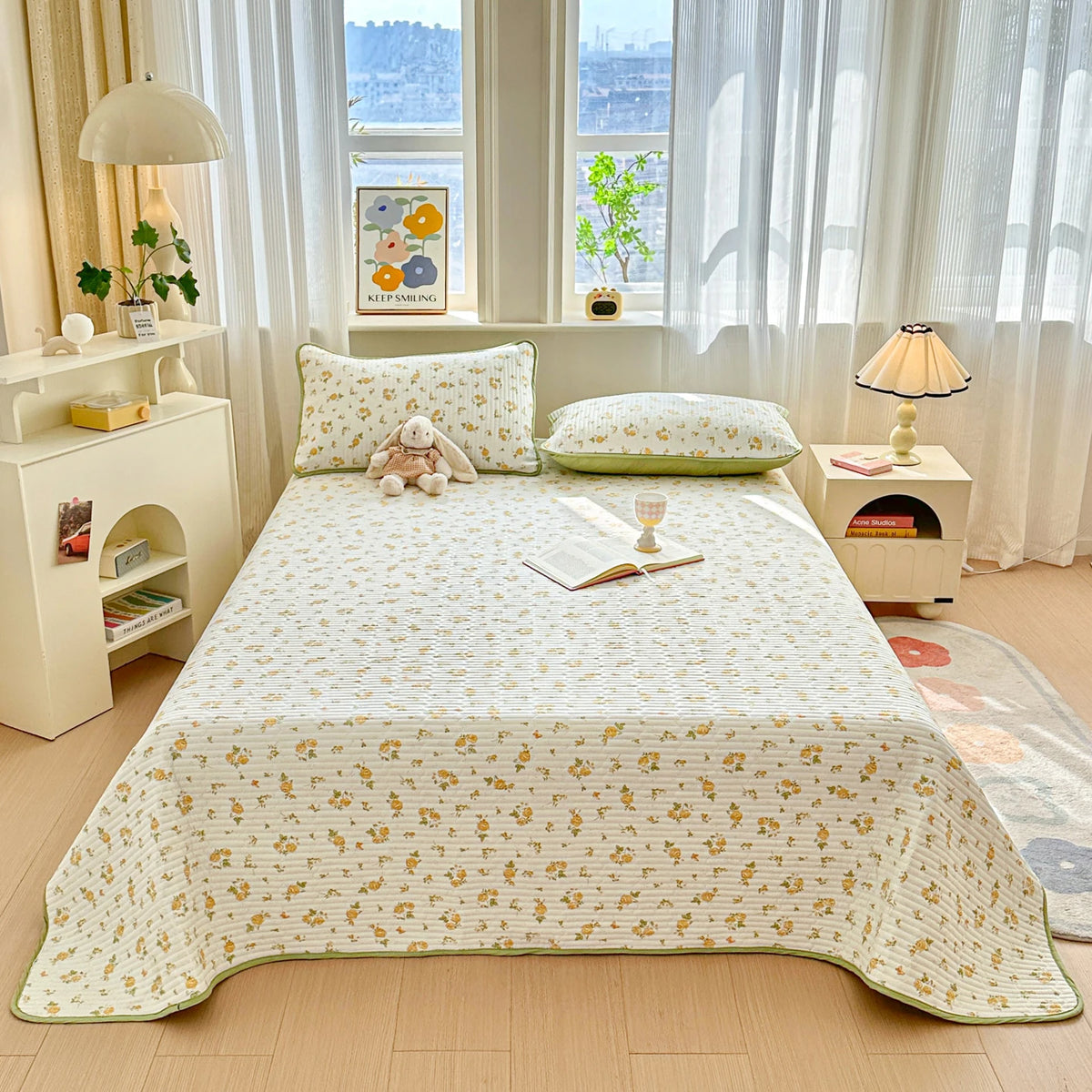 A luxury floral quilted bedspread spread across a light wood bed with white linens and sheer curtains.