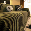 A close-up of a dark olive green herringbone chenille sofa cover with black geometric patterns and white tassel fringes.
