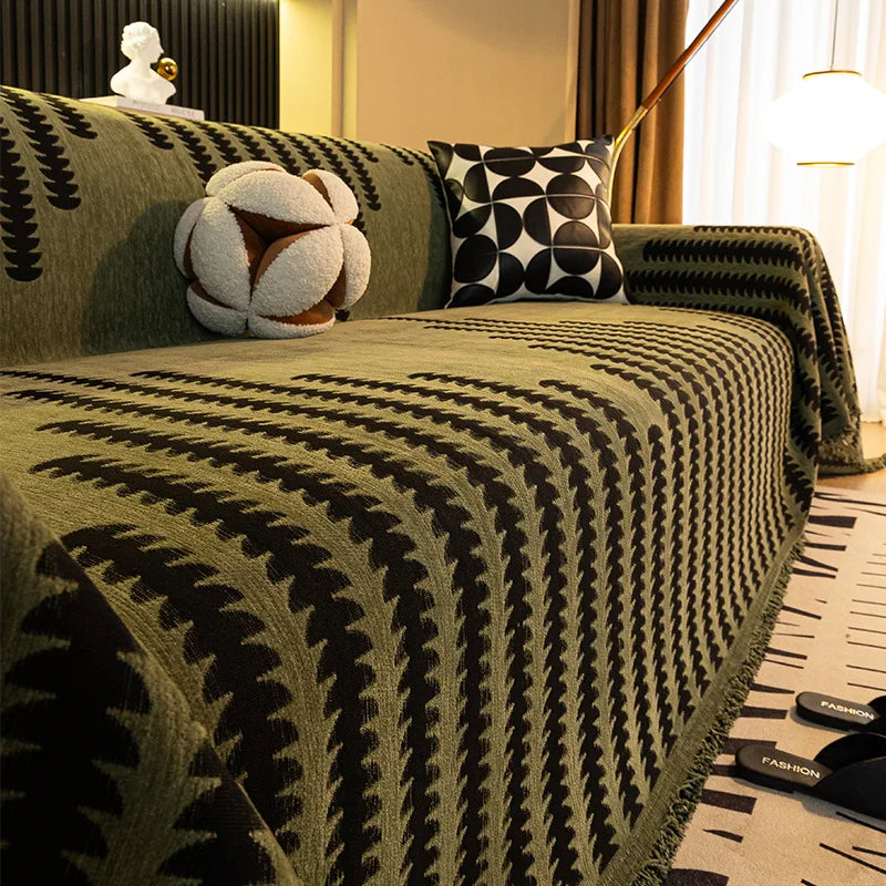 A close-up of a dark olive green herringbone chenille sofa cover with black geometric patterns and white tassel fringes.