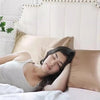 A woman is sleeping comfortably on a bed with a satin pillowcase, resting her head on a large beige pillow.
