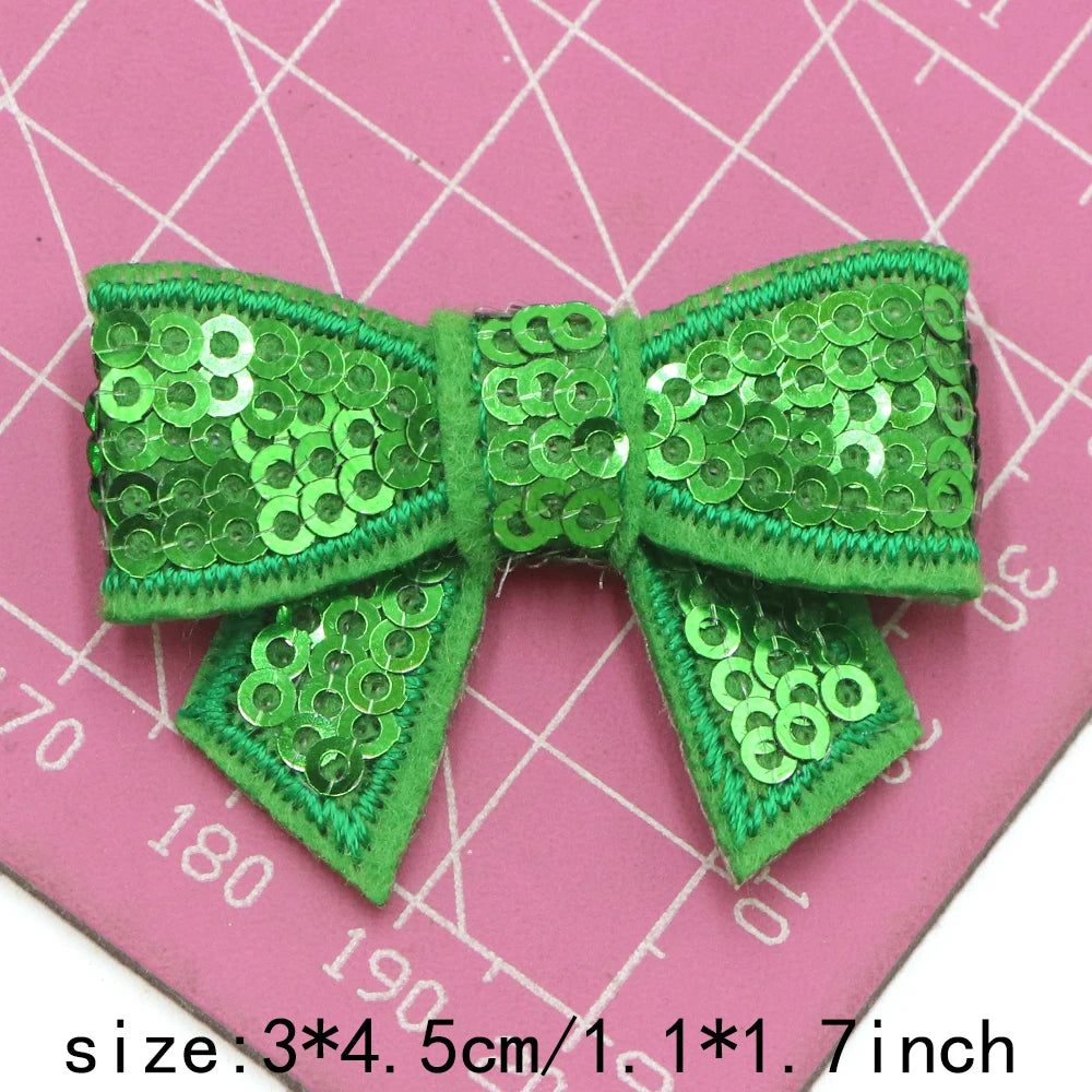 A bright green sequin bow made of fabric is shown against a pink grid background.