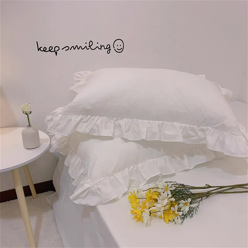 A white ruffled pillow cover is displayed on a white surface with yellow flowers nearby.