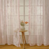 A set of sheer pink floral lace curtains hanging in a window with a small vase of flowers.