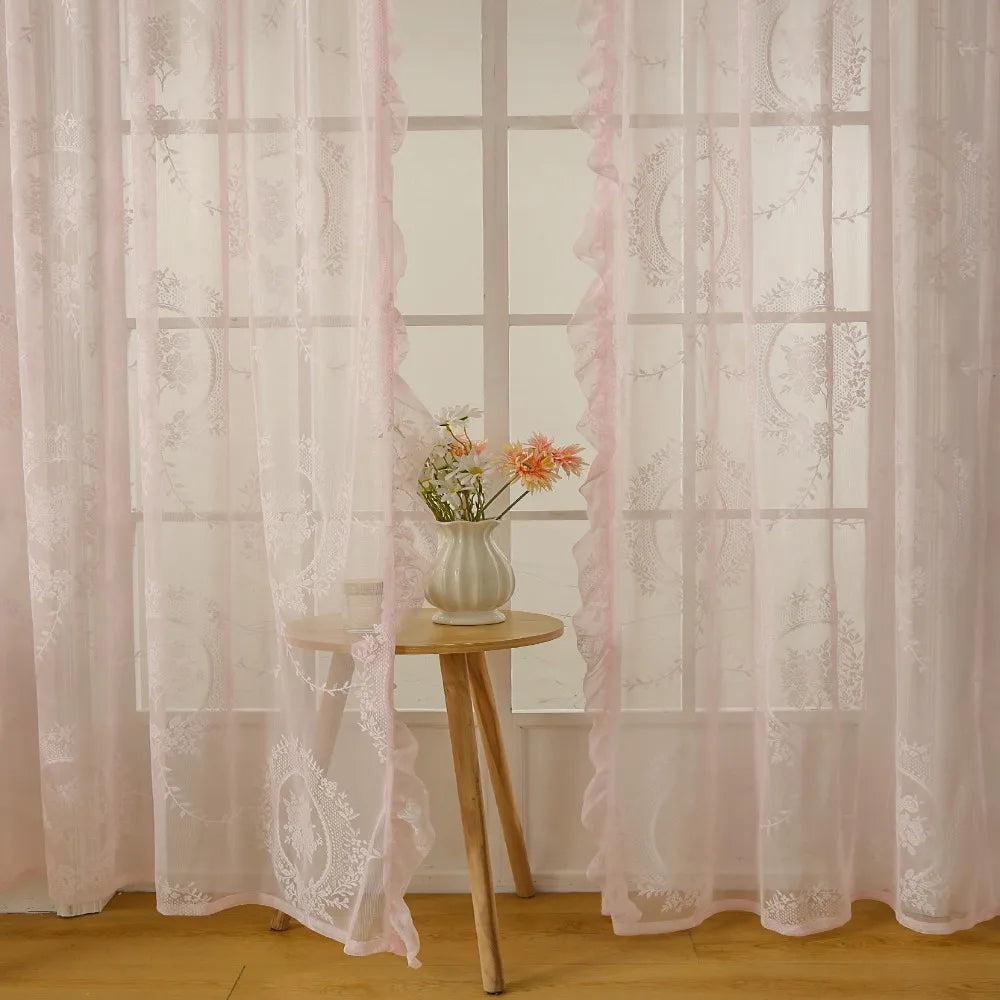 A set of sheer pink floral lace curtains hanging in a window with a small vase of flowers.