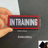 A hand holds a black and red embroidered patch with the text IN TRAINING on it.