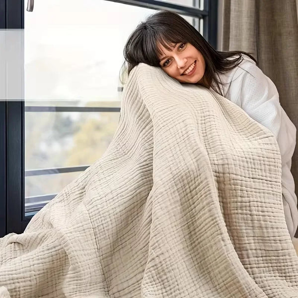 A woman smiling while wrapped in a beige muslin blanket near a window.