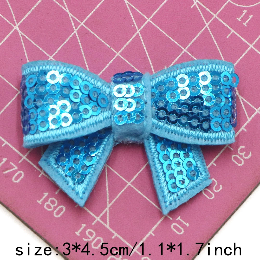 A blue sequin bow made of fabric is shown against a pink grid background.