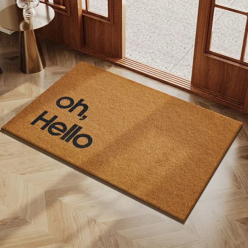 A rectangular, tan coconut fiber doormat with the text oh Hello placed on a light wood floor near a glass door.