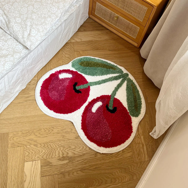 A round, organic-shaped rug featuring embroidered illustrations of red cherries on a light wood floor.