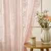 Pink sheer floral lace curtains hanging in a bright room next to a white vase of daisies