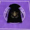 Black velvet pouch with a gold bee emblem resting on a purple surface.