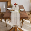 A beige boho hand-woven table runner placed on a wooden dining table with fruit and glassware.