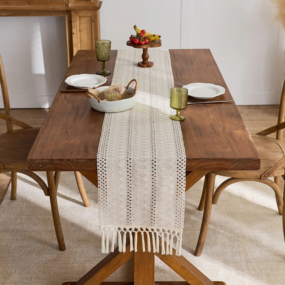 A beige boho hand-woven table runner placed on a wooden dining table with fruit and glassware.
