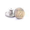 A round brass button with the text BASE GIRL NEW JENS engraved on it, next to its metal finding.