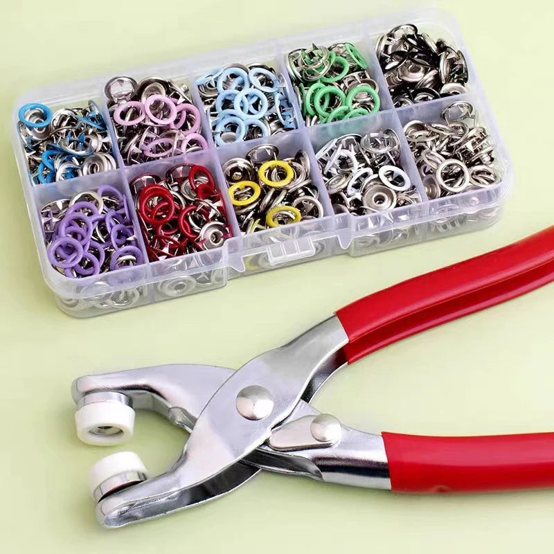 A clear plastic container holds a variety of silver and gold snap buttons, next to a pair of red and silver pliers.