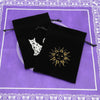 Two black velvet tarot cards storage pouches with white silhouette designs resting on a purple patterned surface.